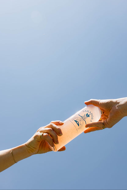 Two hands holding a Sanadora water bottle against a blue sky